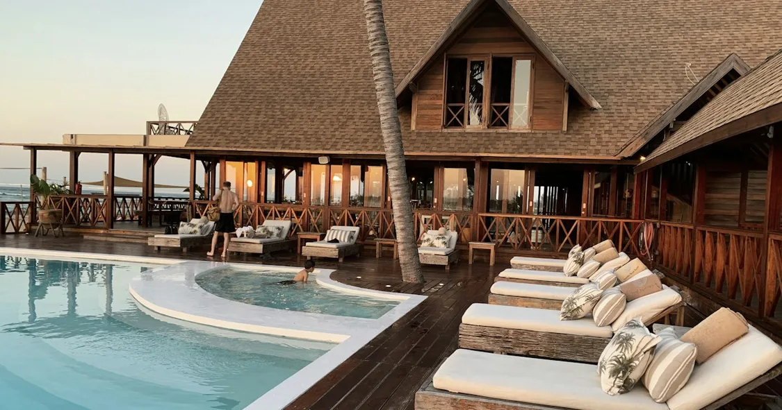 Hotel pool deck with sun loungers in front of a timber-framed resort building at dusk